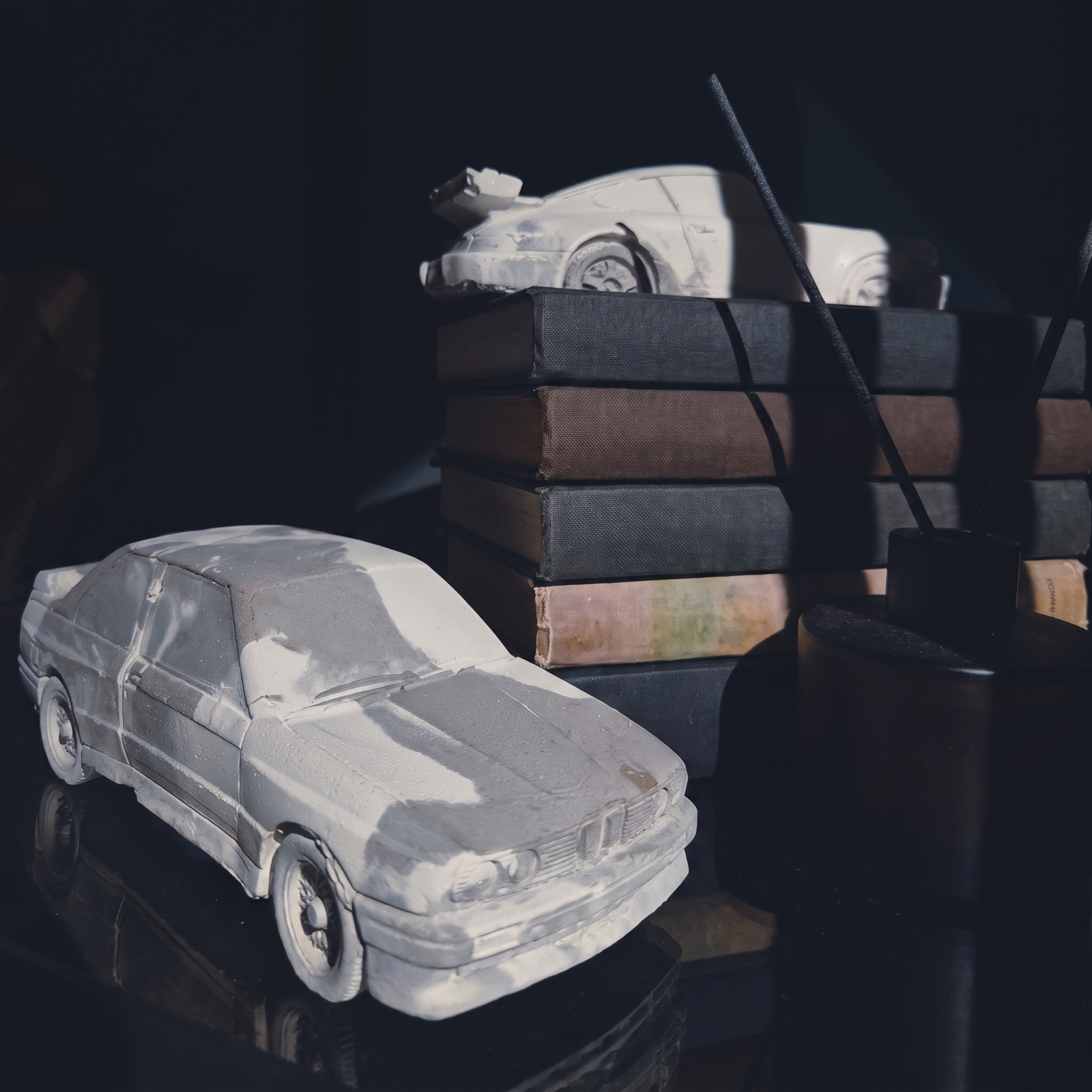 Both Series 01 concrete cars at home on a bookshelf — the saloon on the table, the coupe on a stack of dark books